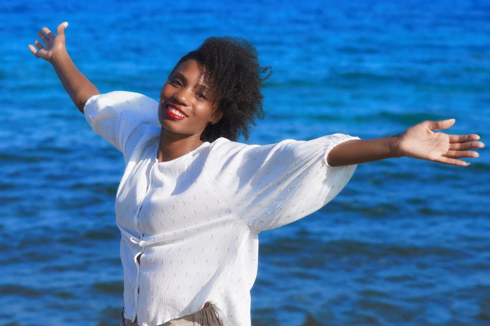 Femme rayonnante aux bras grands ouverts exprimant la joie et la liberté, vêtue de blanc devant un océan bleu azur